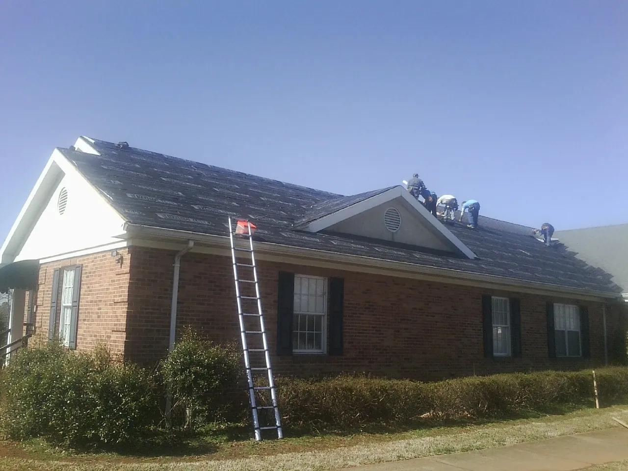 Crew tearing shingles off a brick ranch roof with a ladder set — Metal Roof Repair work by our crew in Peosta
