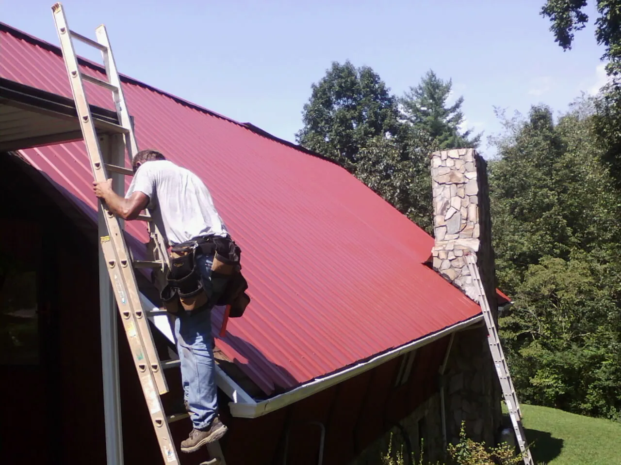 Roofer on a ladder beside a red metal roof and stone chimney — Soffit & Fascia Repair by Roofing Brothers in Peosta